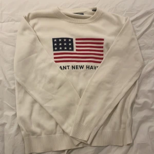 Vit Gant tröja med USA-flagga - Vit långärmad tröja från Gant med broderad amerikansk flagga och texten 'GANT NEW HAVEN' på bröstet. Tröjan har ribbade muddar och är tillverkad i mjuk bomull. Perfekt för dig som gillar klassisk och stilren college-look.