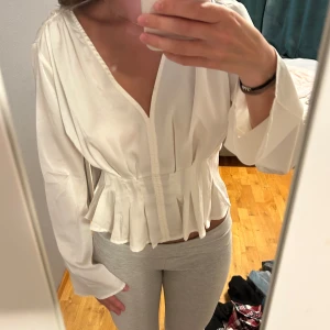 Vit omlottblus med volangdetalj - Säljer en superfin vit blus med v-ringning och omlottkänsla. Blusen har långa ärmar och markerad midja med volangdetaljer nedtill som ger en snygg siluett. Perfekt för dig som gillar en stilren men ändå trendig look.