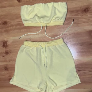 Gult matchande set från Femme Luxe - Snygga ljusgula shorts från Femme Luxe med elastisk midja och snörning framtill. Mjukt och lätt material som känns skönt mot huden, perfekt för varma dagar. Matchande bandeau-topp ingår. Shortsen har en avslappnad passform och sportig vibe.