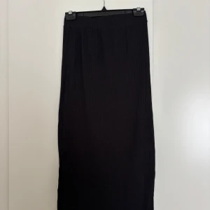 Svart ribbad långkjol från Pieces - Svart långkjol från Pieces i ribbad trikå med slits på sidan. Kjolen har en enkel och clean look som är lätt att styla med allt. Perfekt för dig som gillar minimalistiska vibes och vill ha något bekvämt men ändå snyggt.