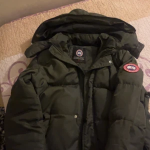 Jacka  Canada Goose - Säljer en grön dunjacka från Canada Goose med huva och klassisk logga på ärmen. Jackan är tjock och varm, perfekt för kalla dagar. Stängs med dragkedja och knappar framtill. Snygg och stilren design som håller dig varm hela vintern.
