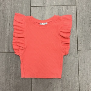Koralldröd ribbad topp från Zara - Superfin korallröd topp från Zara med ribbad struktur och volangärmar. Modellen är croppad och har en rund halsringning. Perfekt för dig som vill ha en färgklick i garderoben och gillar trendiga detaljer.