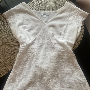 Vit tie topp - Superfin vit tie topp i bomull med broderade blommor och v-ringning. Toppen har korta ärmar och knytband i midjan för en snygg passform. Perfekt för sommarens alla tillfällen! Det är storlek S! Har bara använt den 1 gång!