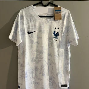 Frankrike fotbollströja Nike vit - Snygg vit Frankrike fotbollströja från Nike med diskret mönster av franska landmärken och blå tupp-logga med två stjärnor på bröstet. Kortärmad modell i lätt och ventilerande Dri-FIT-material, perfekt för match eller träning.