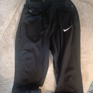Svarta Nike Dri-FIT träningsbyxor - Svarta träningsbyxor från Nike med Dri-FIT-teknologi och vit swoosh-logga på låret. Byxorna har resår i midjan och smal passform, perfekta för gymmet eller löprundan. Snygg och sportig look med praktiska fickor på sidorna.