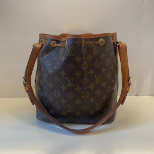 Louis Vuitton Monogram Noé axelväska - Säljer en klassisk Louis Vuitton Noé axelväska i brunt monogrammönster med justerbar axelrem och snörning upptill. Väskan är tillverkad i skinn och canvas med detaljer i läder. Rymlig insida och ikonisk LV-logga över hela väskan.