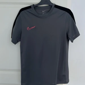 Grå Nike t-shirt med rosa logo - Snygg grå t-shirt från Nike med korta ärmar och rosa swoosh på bröstet.