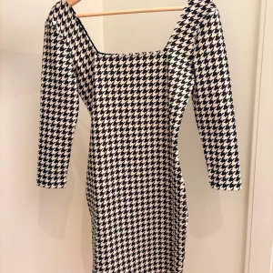 Chic Svartvit klänning från Easy Wear - Snygg svartvit klänning från Easy Wear i klassiskt hundtandsmönster. Modellen är figurnära med trekvartsärm och fyrkantig ringning. Perfekt för dig som vill sticka ut med en tidlös look.