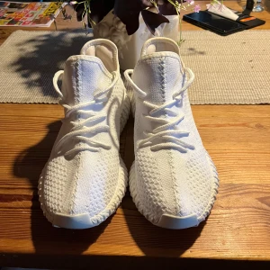 Vita Adidas Yeezy Boost sneakers - Säljer ett par helvita Adidas Yeezy Boost sneakers med stickad ovandel och snörning. Skorna har en chunky sula och klassisk Yeezy-design, perfekt för dig som gillar streetstyle. Materialet är syntet och mesh för en luftig känsla.