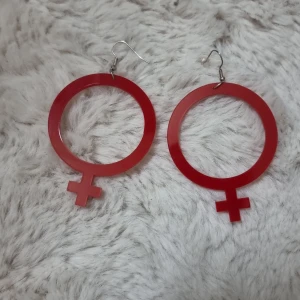 Stora röda örhängen kvinnosymbol - Statementörhängen i form av kvinnosymboler, tillverkade i röd plast. De är stora och runda med ett tydligt kors nedtill, och har silvriga krokar för enkel användning. Väldigt lätta och bekväma att ha på sig. Sparsamt använda.