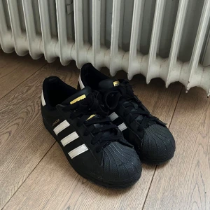 Adidas Superstar svarta sneakers - Klassiska Adidas Superstar sneakers i svart med vita ränder och gulddetaljer på plösen. Använda men i mycket bra skick! Nypris på 1400kr och säljer för 400🩷