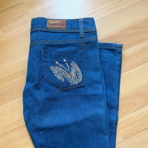 Blå bootcut jeans med broderad ficka - Snygga blå jeans från Leadoff med bootcut passform och låg midja. Bakfickan har ett coolt broderi i flera färger som ger extra edge. Klassisk denim med orange sömmar och bälteshällor. .