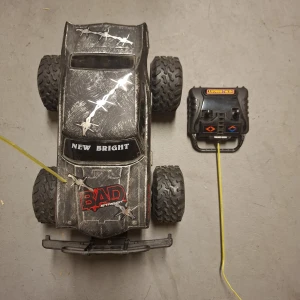 Radiostyrd bil - Bilderna visar en radiostyrd bil av modell "New Bright Bad Street" med tillhörande fjärrkontroll. Bilen har stora terrängdäck och en cool, sliten design med taggtrådsdekor. Perfekt för dig som gillar fartfyllda utomhuslekar och vill ha en robust leksaksbil att köra med vänner.