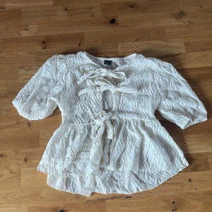 Vit blus med knyt från Gina Tricot - Superfin vit blus från Gina Tricot med crinklad struktur och tre knyt framtill. Blusen har volangig nederdel och halvlånga puffärmar. 