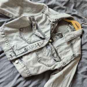 Ljusgrå jeansjacka från Gina Tricot - Snygg ljusgrå jeansjacka från Gina Tricot med klassisk krage, knappar framtill och två bröstfickor. Jackan har en croppad passform och markerade sömmar som ger en cool vintagekänsla. Perfekt att slänga över en hoodie eller t-shirt.