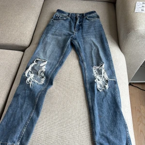 Blå ripped jeans från NA-KD, strl 34 - Säljer ett par blå jeans från NA-KD i storlek 34. De har raka ben och stora slitningar med trasiga hål på båda knäna för en riktigt edgy look. Klassisk femficksmodell och hög midja. Perfekta för dig som gillar streetstyle.