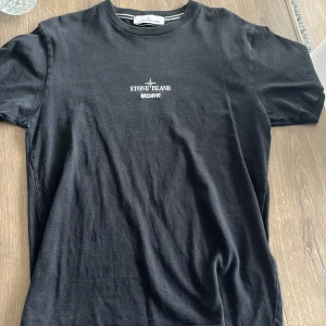 Svart Stone Island t-shirt med tryck - Svart t-shirt från Stone Island med liten vit logga och ARCHIVIO-tryck på bröstet. Baksidan har ett stort tryck med jacka och text. Klassisk rund hals och korta ärmar. Skön passform och tillverkad i mjuk bomull, perfekt för en clean streetwear-look. Ny pris 2500 och passar även M