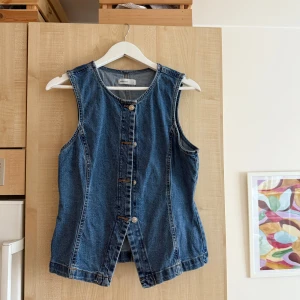 Jeansväst från GinaTricot - Snygg jeansväst från GinaTricot i klassisk blå denim. Västen har rund halsringning, knäppning framtill med silverfärgade knappar och coolt snitt nertill. Perfekt att styla över en t-shirt eller hoodie för en avslappnad vibe.
