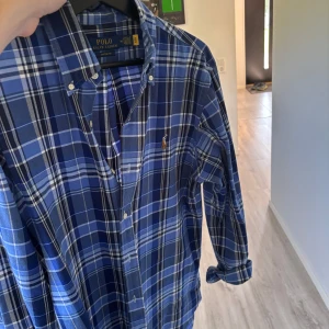 Skjorta Polo Ralph Lauren M - Snygg blårutig skjorta från Polo Ralph Lauren i custom fit. Skjortan har klassisk krage, knappar framtill och lång ärm. Ikoniska broderade loggan på bröstet. Perfekt för dig som gillar stilren och tidlös design.