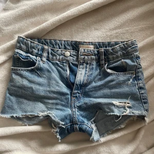 Blå jeansshorts från Gina Tricot - Snygga blå jeansshorts från Gina Tricot x Izaandelle med råa, fransiga kanter och slitna detaljer framtill. Klassisk femficksmodell med knapp och dragkedja. Perfekta för varma dagar och en avslappnad look. Säljer pga för små för mig!