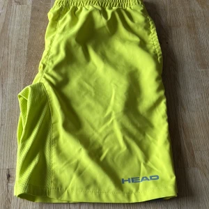 Neongula träningsshorts från HEAD - Snygga neongula träningsshorts från HEAD med elastisk midja och ventilerande meshpanel på sidan. Perfekta för dig som vill sticka ut på tennisbanan eller gymmet. Lätta och bekväma i syntetmaterial som andas. Logga i blått framtill.