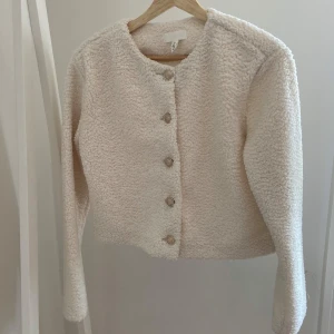 Beige teddyjacka från H&M - Supermjuk och fluffig teddyjacka från H&M i beige. Jackan har rund halsringning, långa ärmar och snygga silverfärgade knappar framtill. Perfekt för lager-på-lager och ger en chill vibe till din outfit. Använd 1 gång