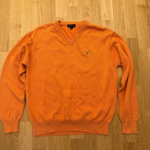 Orange v-ringad tröja från Gant - Snygg orange v-ringad tröja från Gant i mjuk bomull. Klassisk passform med ribbade muddar och diskret broderad logga på bröstet. Perfekt för dig som vill sticka ut med färg och ändå hålla det stilrent.
