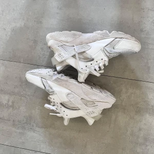 Vita Nike Air Huarache sneakers - Säljer ett par vita Nike Air Huarache sneakers med chunky sula och klassisk design. Skorna har snörning, rund tå och detaljer i syntet och skinn. Perfekta för dig som gillar en sportig och clean stil.