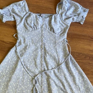 Ljusblå blommig klänning Bubbleroom - Supersöt ljusblå kortklänning från Bubbleroom med små vita blommor, puffärmar och knappar framtill. Den har en knytdetalj som funkar fram & baktill