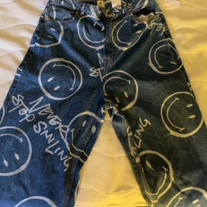 Y2K Blå jeans med smiley-mönster - Unika blå jeans med stora vita smiley-ansikten och texten 'Never Stop Smiling' tryckt över hela byxorna. Klassisk femficksmodell i denim med normal passform. Perfekta för dig som vill sticka ut och sprida good vibes.