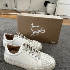 Vita sneakers från Christian Louboutin - Snygga vita sneakers från Christian Louboutin med ikoniska röda sulor och coola nitar framtill. 