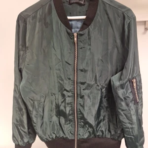 Mörkgrön bomberjacka från Zanzea - Snygg mörkgrön bomberjacka från Zanzea med svarta ribbade muddar och dragkedja framtill. Jackan har en klassisk ärmficka med dragkedja och en glansig finish. Perfekt för dig som vill ha en stilren och trendig look.