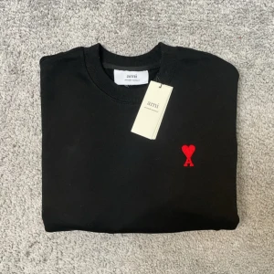 Svart sweatshirt från Ami Paris - Svart sweatshirt från Ami Paris med klassisk rund hals och lång ärm. På bröstet finns det ikoniska röda Ami de Coeur-broderiet. Tröjan är i mjuk bomull och har ribbade muddar vid ärmslut och nederkant. Perfekt för en clean och stilren look. Pris kan diskuteras!