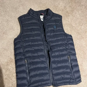 Mörkblå dunväst från U.S. Polo Assn. -    mörkblå dunväst från U.S. Polo Assn. med quiltad design och broderad logga på bröstet. Västen har två dragkedjeförsedda fickor och hög krage. Perfekt att slänga över en hoodie för extra värme. Storlek 14-15 år. Skulle säga att den passar xs-s. 