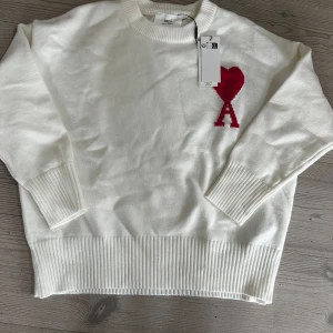 Vit stickad tröja från Ami paris S - Säljer en stilren vit stickad tröja från Ami Paris med ett rött hjärta och bokstaven A på bröstet. Tröjan har långa ärmar och ribbade muddar vid ärmslut och nederkant. Perfekt för en trendig och bekväm look.