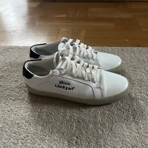 Vita sneakers från Saint Laurent - Snygga vita sneakers från Saint Laurent med svart häl och handskriven logga på sidan. Skorna har klassisk snörning och en stilren design som passar perfekt för en trendig look.