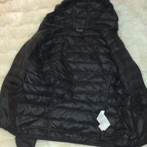 Svart pufferjacka från Emporio Armani - Snygg svart pufferjacka från Emporio Armani med guldfärgad logga på bröstet. Jackan har en dragkedja framtill och en praktisk huva. Perfekt för kyligare dagar och ger en stilren look.