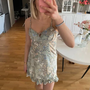 Mönstrad kortklänning med volangdetaljer - Säljer en söt kortklänning med ett färgglatt mönster. Klänningen har smala axelband och en volangkant längst ner. 