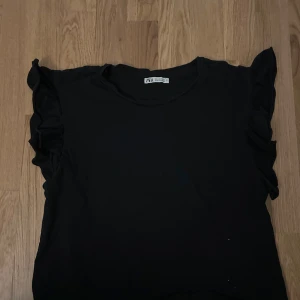 Svart topp med volangärmar från Zara - Snygg svart topp från Zara med volangdetaljer på ärmarna. Perfekt för en stilren look. Toppen har en enkel design som gör den lätt att matcha med olika outfits.