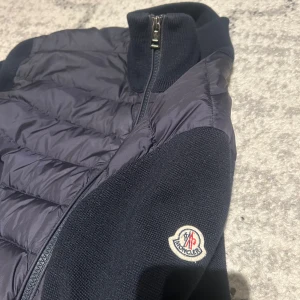 Mörkblå jacka från Moncler - Snygg mörkblå jacka från Moncler med quiltad framsida och stickade ärmar. Jackan har en dragkedja framtill och en hög krage för extra värme. Perfekt för kyliga dagar. Skick 8/10 då den har ett sytt ”jack” som bild 5 visar.