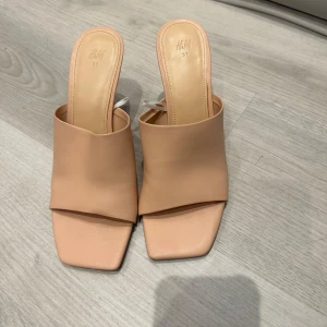 Beige klackskor från H&M - Snygga beige klackskor från H&M med öppen tå och fyrkantig form. Skorna har en stilren design med en bred rem över foten och en bekväm klack. Perfekta för en elegant look.
