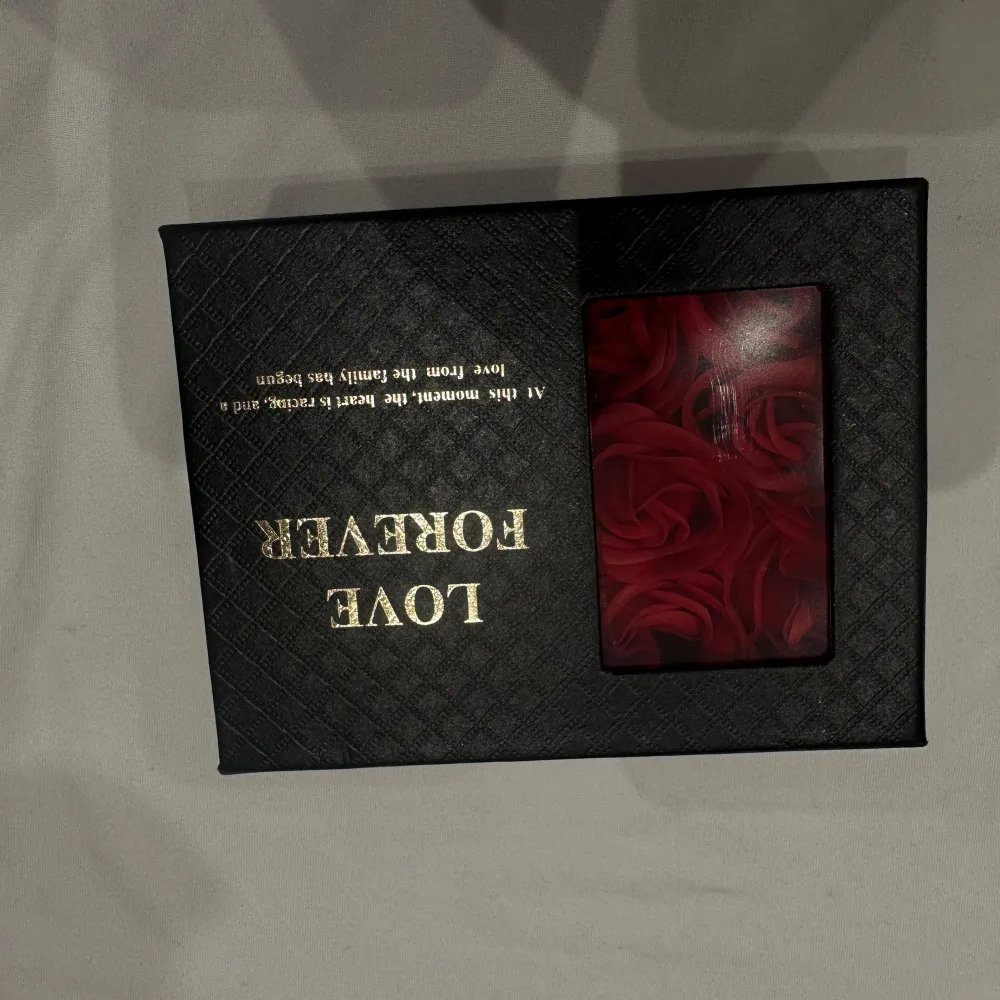Elegant presentask med konstgjorda röda rosor. Asken är svart med texten 'Love Forever' i guld och har ett fönster som visar rosorna. Perfekt som en romantisk gåva eller dekoration.. Asusteet.