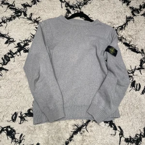 Grå stickad tröja från Stone Island - Säljer en stilren grå stickad tröja från Stone Island. Tröjan har långa ärmar och en rund halsringning. Den ikoniska logotypen sitter på ärmen. Perfekt för en avslappnad och trendig look.