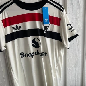 Vit och röd fotbollströja från Adidas - Snygg vit fotbollströja från Adidas med röda och svarta ränder över bröstet. Tröjan har korta ärmar och tryck på både fram- och baksidan. Perfekt för fotbollsfans som vill visa sitt stöd.