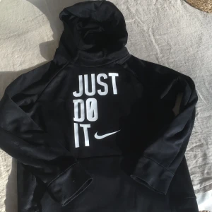 Svart hoodie från Nike - Säljer en svart hoodie från Nike med den ikoniska texten 'Just Do It' i vitt på framsidan. Hoodien är tillverkad i Dri-FIT-material för att hålla dig torr och bekväm. Perfekt för träning eller vardagsbruk.