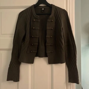 Olivgrön cardigan jacka från H&M - Säljer en supersnygg olivgrön cardigan jacka från H&M med dekorativa knappar framtill. Jackan har en kort/medellång passform och långa ärmar med ribbade detaljer. 