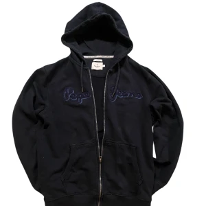Mörk blå hoddie från Pepe Jeans - Säljer en svart hoodie från Pepe Jeans med dragkedja och broderad logga på bröstet. Den har en justerbar huva med snören och praktiska fickor framtill. Perfekt för en avslappnad stil.