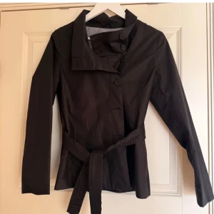 Svart kavaj med knyt skärp - Snygg svart kavaj med knappar och bälte i midjan. Perfekt för en stilren look. Långärmad och med en elegant krage.