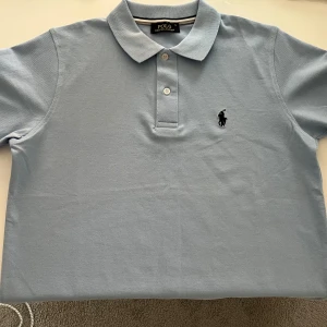Ljusblå pikétröja från Polo Ralph Lauren - Säljer en klassisk ljusblå pikétröja från Polo Ralph Lauren med den ikoniska loggan broderad på bröstet. Tröjan har en krage och knappar framtill. Perfekt för en stilren och avslappnad look. Aldrig använd, nyskick.