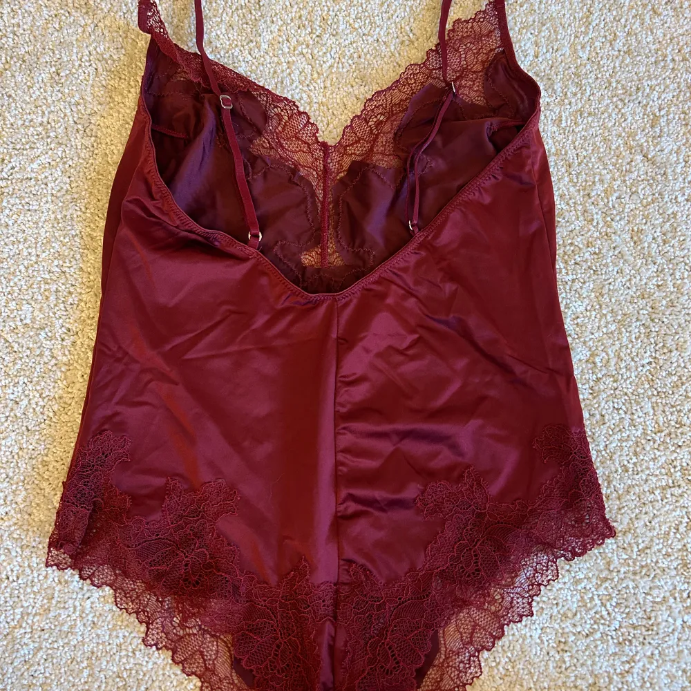 Elegant vinröd body med spetsdetaljer och justerbara axelband. Den har en djup v-ringning och spetskant runt benöppningarna för en feminin touch. Den är aldrig använd endast testad🥰. Yläosat.